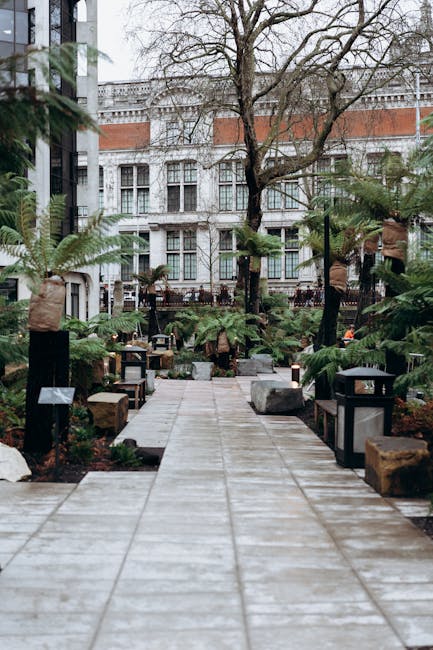 A paved walkway leading through a lush, well-maintained garden courtyard in front of a historic white stone building with large windows. The garden features various plants, including ferns, small shru