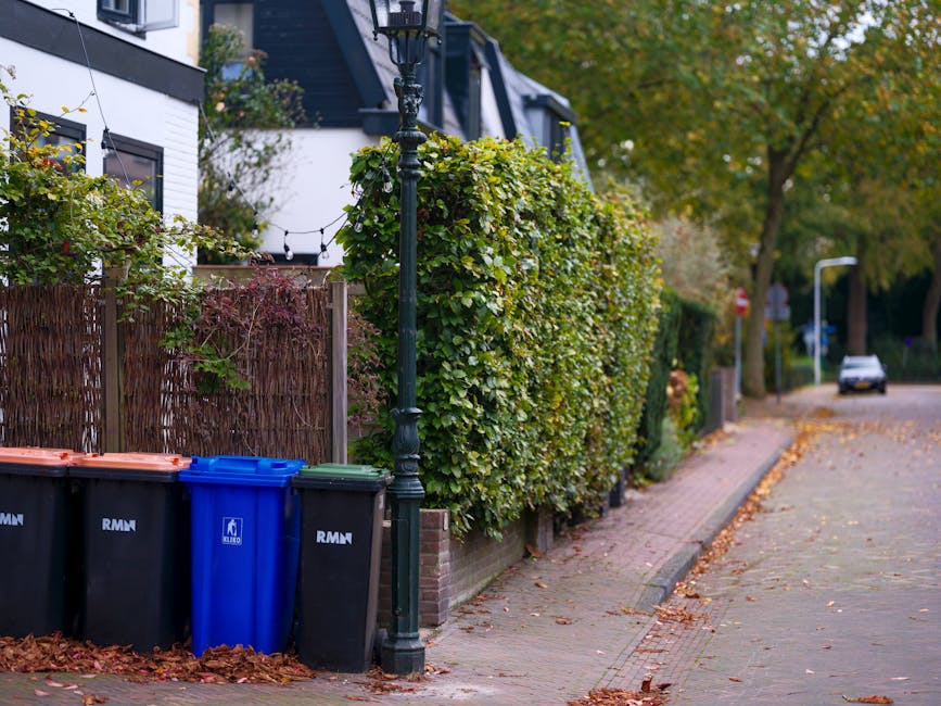 A residential street scene featuring a row of three large wheelie bins—a black, a blue, and a dark green—placed near a black metal lamppost on the edge of a sidewalk. Behind the bins, there is a t